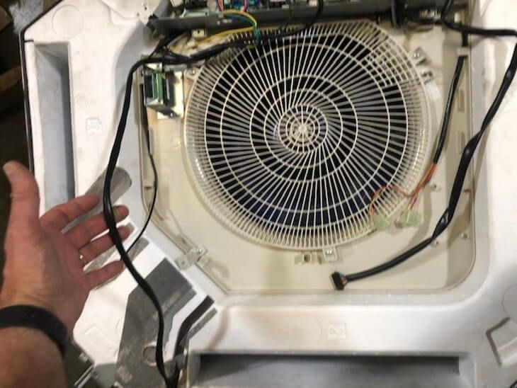 Hand beside a circular fan assembly inside a white appliance compartment with exposed wiring.