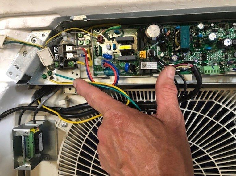 Hand pointing to wiring and circuit board inside an HVAC unit near a fan