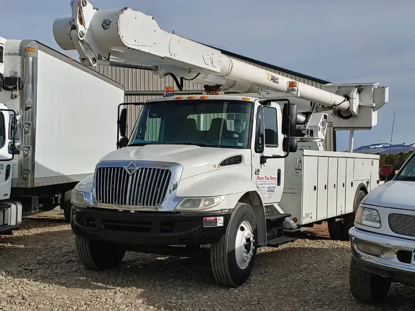 A white utility truck with a crane on top of it is parked in a gravel lot.