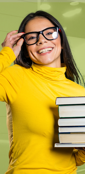 Una mujer que lleva gafas y un suéter amarillo sostiene una pila de libros.