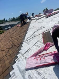 Roofers installing white underlayment on a sloped roof beside brown shingles under a clear sky