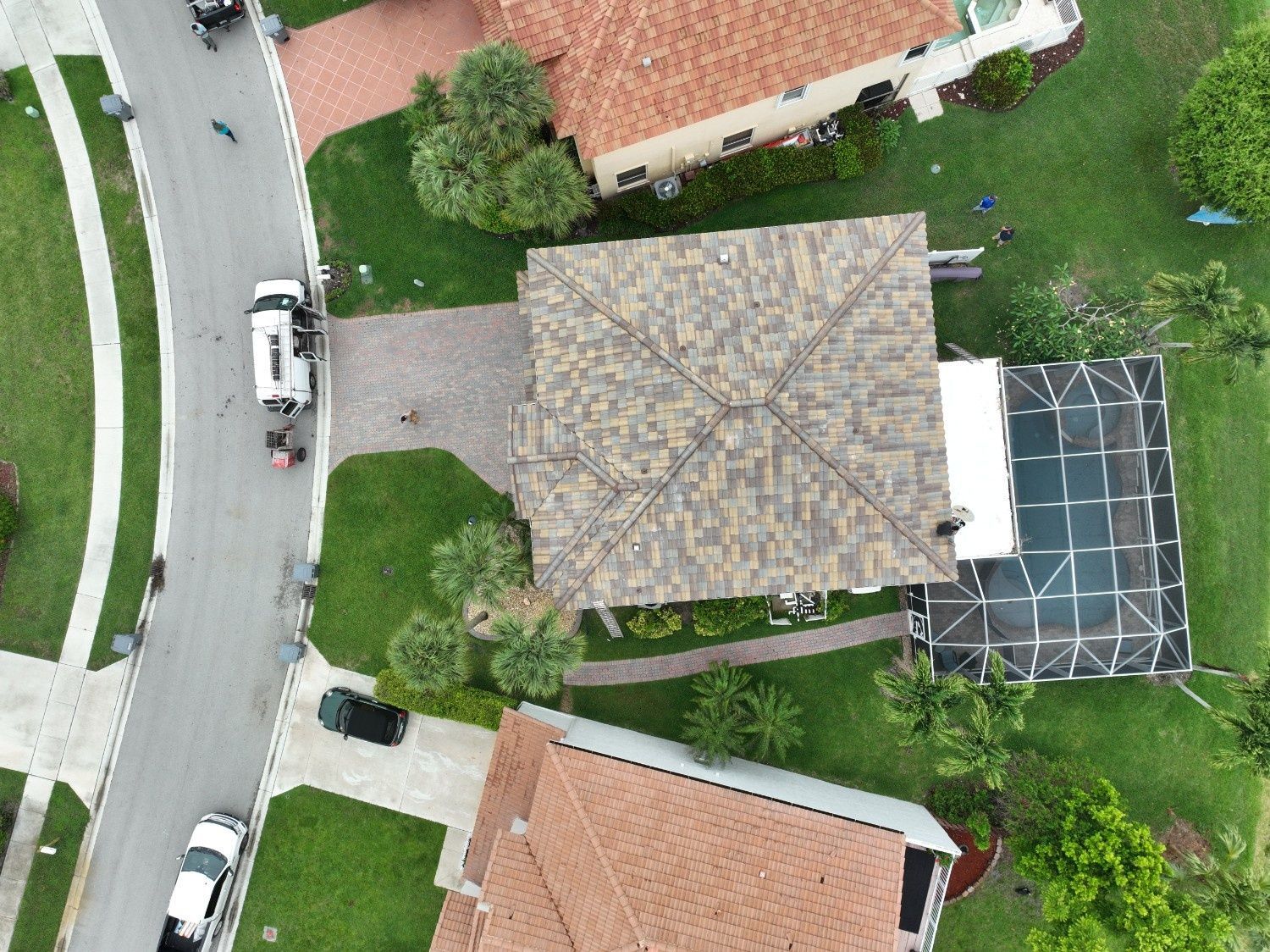 Aerial view of a house with a gray roof, driveway, parked cars, and surrounding trees.