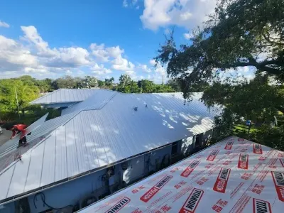 Workers installing a metal roof on a large building under a bright blue sky