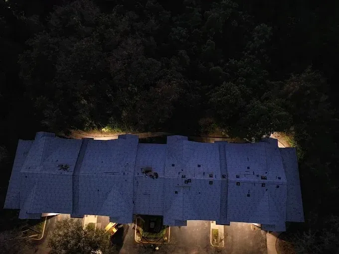 Aerial view of a dark-roofed house surrounded by trees, lit by warm lights at night.