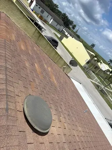 Angled view of a red shingle roof with a round vent, facing a residential street and houses under a cloudy sky