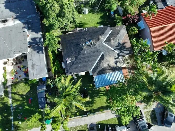 Aerial view of a house with a dark roof, surrounded by lush greenery, neighboring homes, and a blue awning.