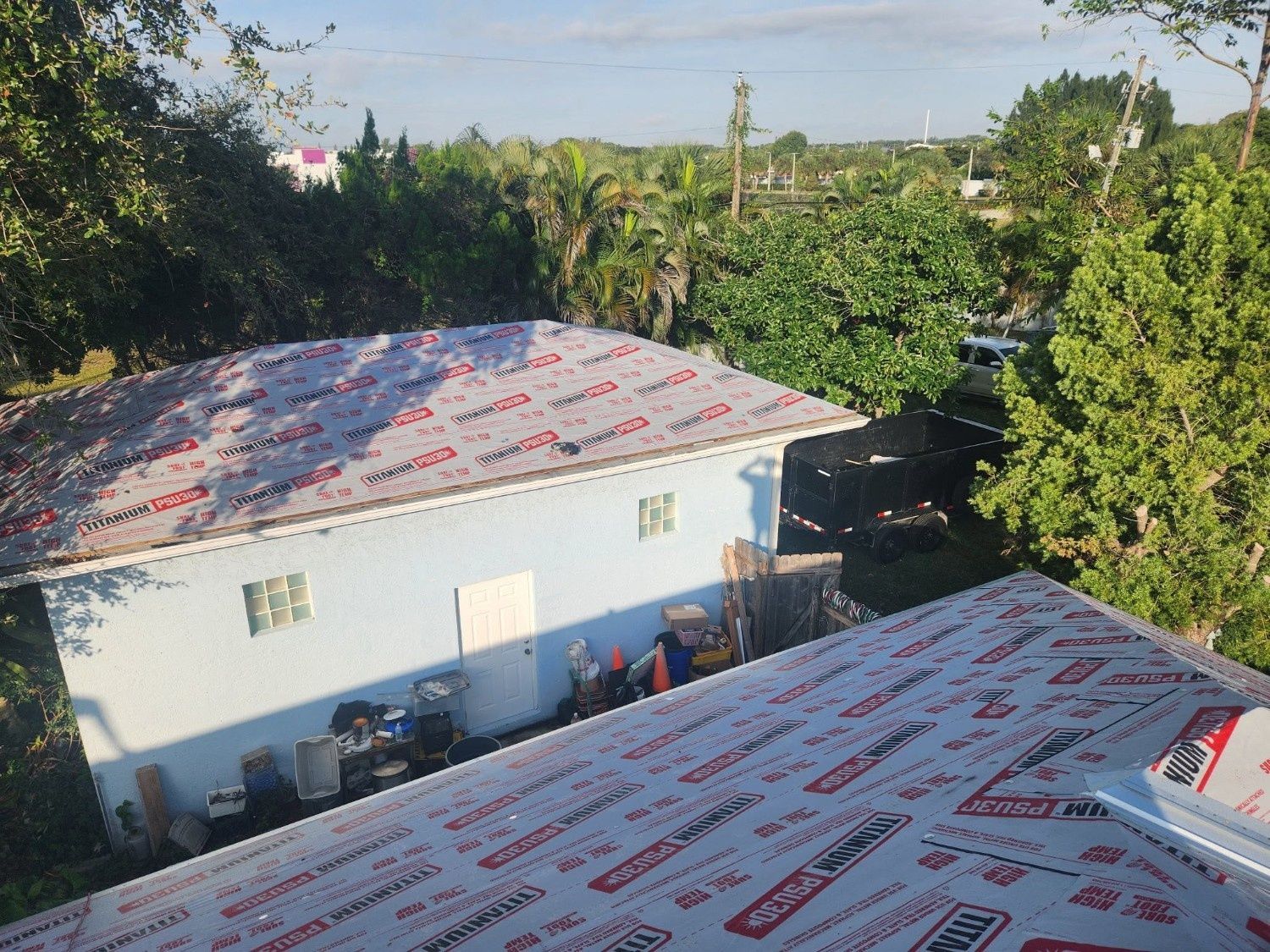 Rooftops with protective wrap, a white house, and lush green trees under a sunny sky.
