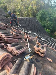 Worker securing damaged roof tiles with ropes on a sloped rooftop amid trees