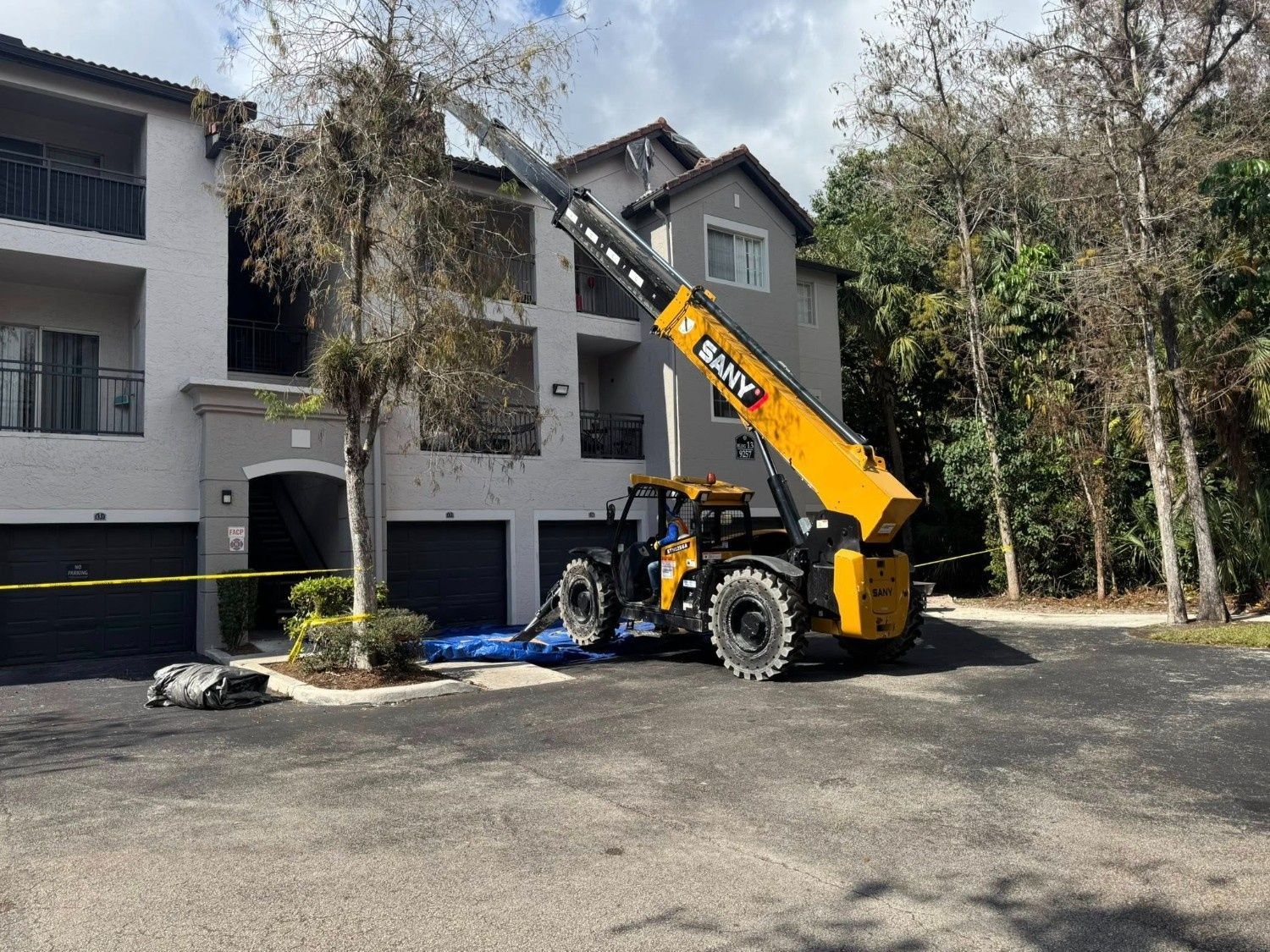 Yellow crane lifting a tree in front of a gray house, with workers and debris on the driveway