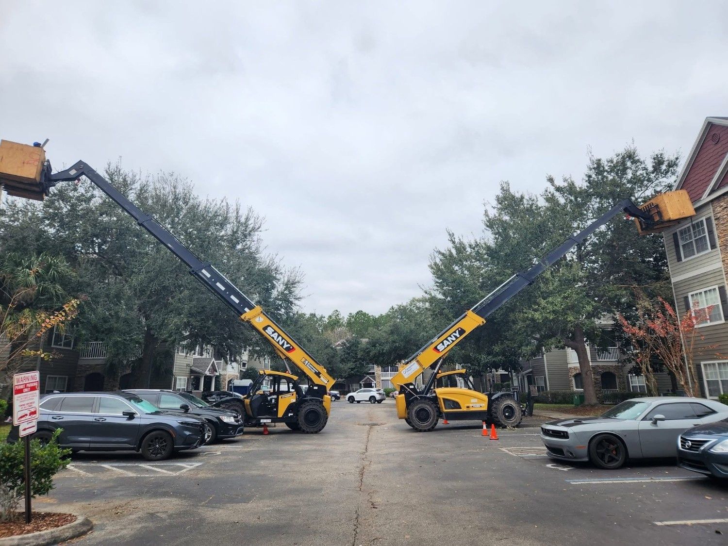 Two yellow cranes raising long booms over a street lined with parked cars and trees