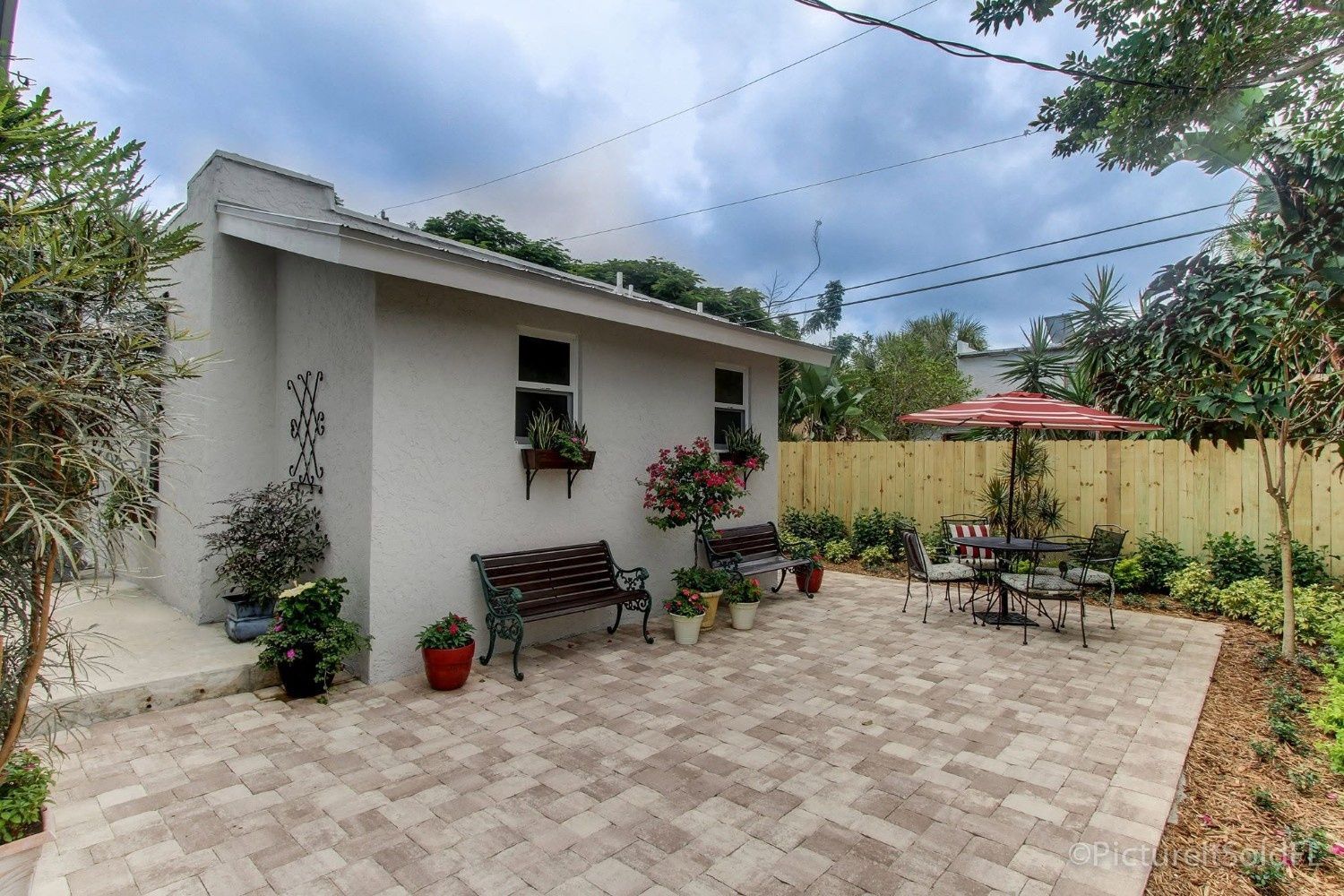 Small white garden cottage with patio seating, potted plants, and a paved courtyard under a cloudy sky