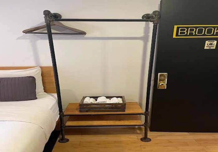 Small hotel-style clothes rack with wooden shelves beside a bed and black door.