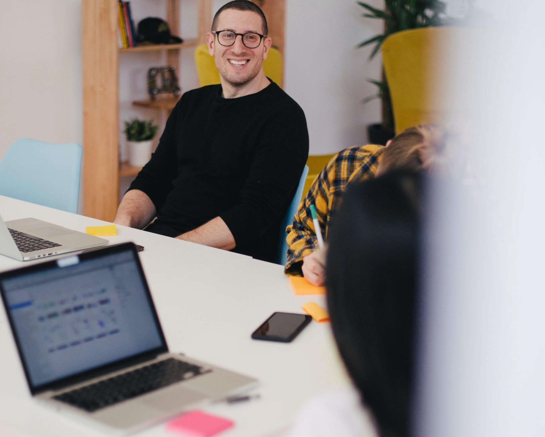 Smiling man in black sweater at a conference table with laptops in a bright office