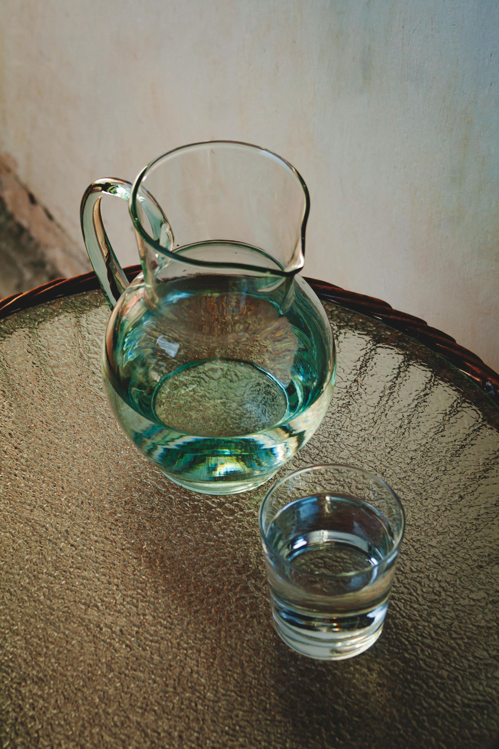 Glass pitcher and small tumbler on a tabletop against a plain wall.