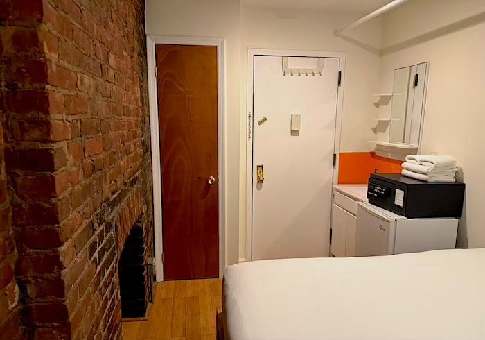 Small room with exposed brick wall, wooden door, white cabinets, and a bed in the foreground.