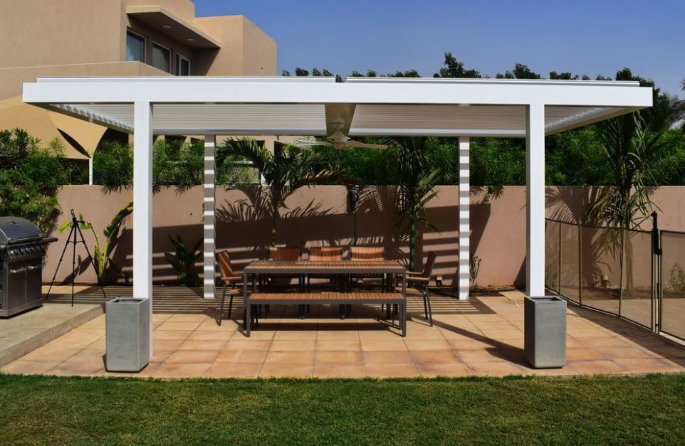 Patio with white pergola over a wooden table and benches. Green grass and a grill in the background.