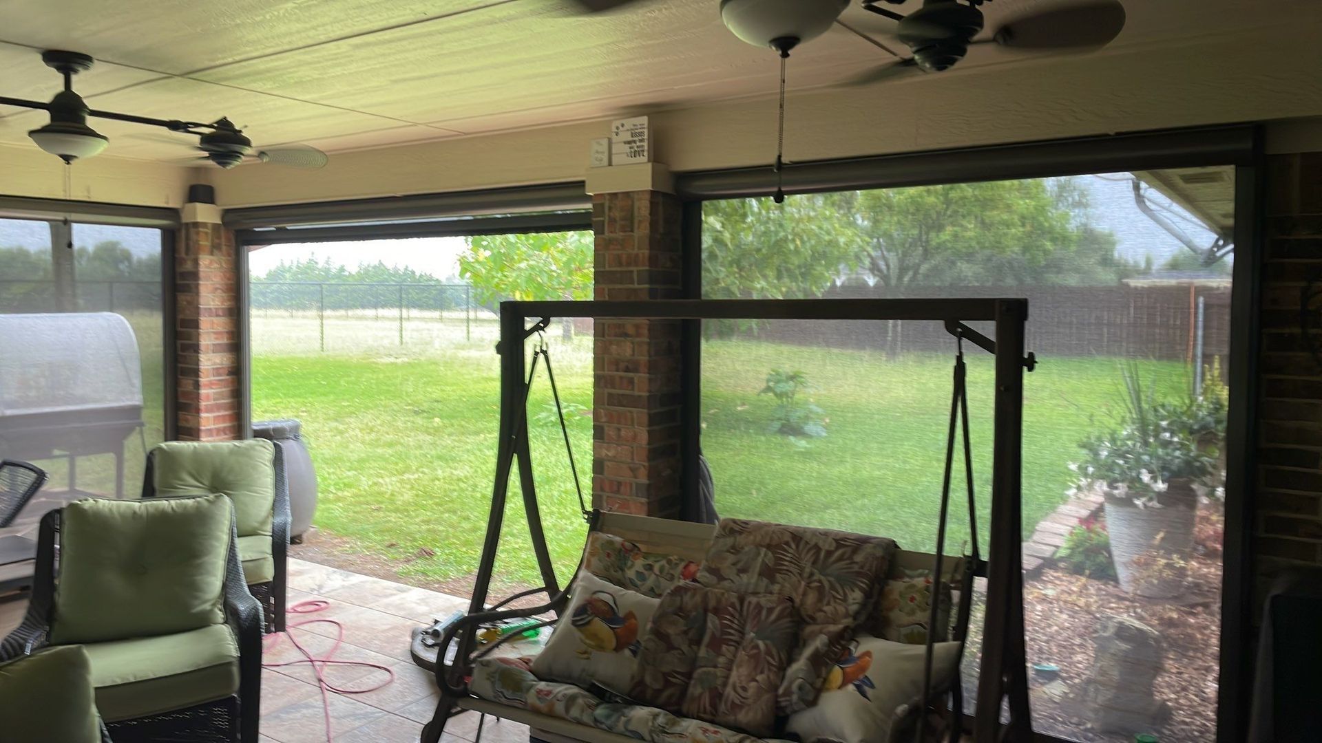 Covered patio with retractable shades, overlooking a grassy field.  Swing, chairs, and brick columns are visible.