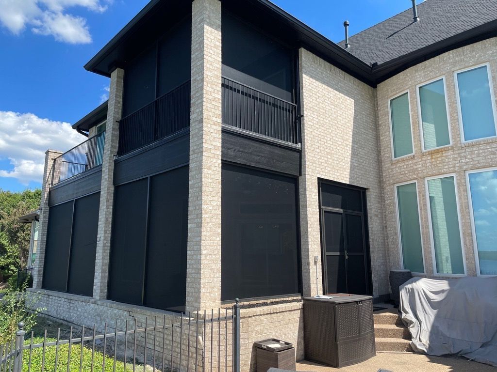 Black retractable sun screens installed on a two-story brick home's patio area, with a black balcony railing.