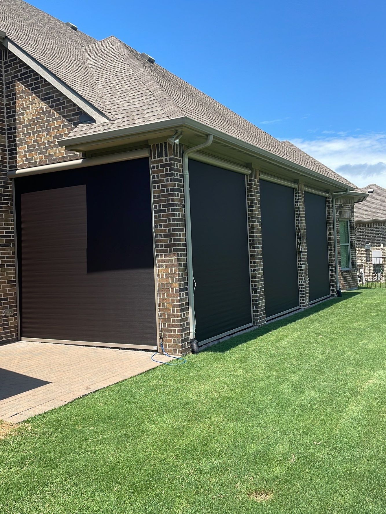 Exterior of a house with brown siding and brick columns; three black shades are lowered on the patio, on green grass.