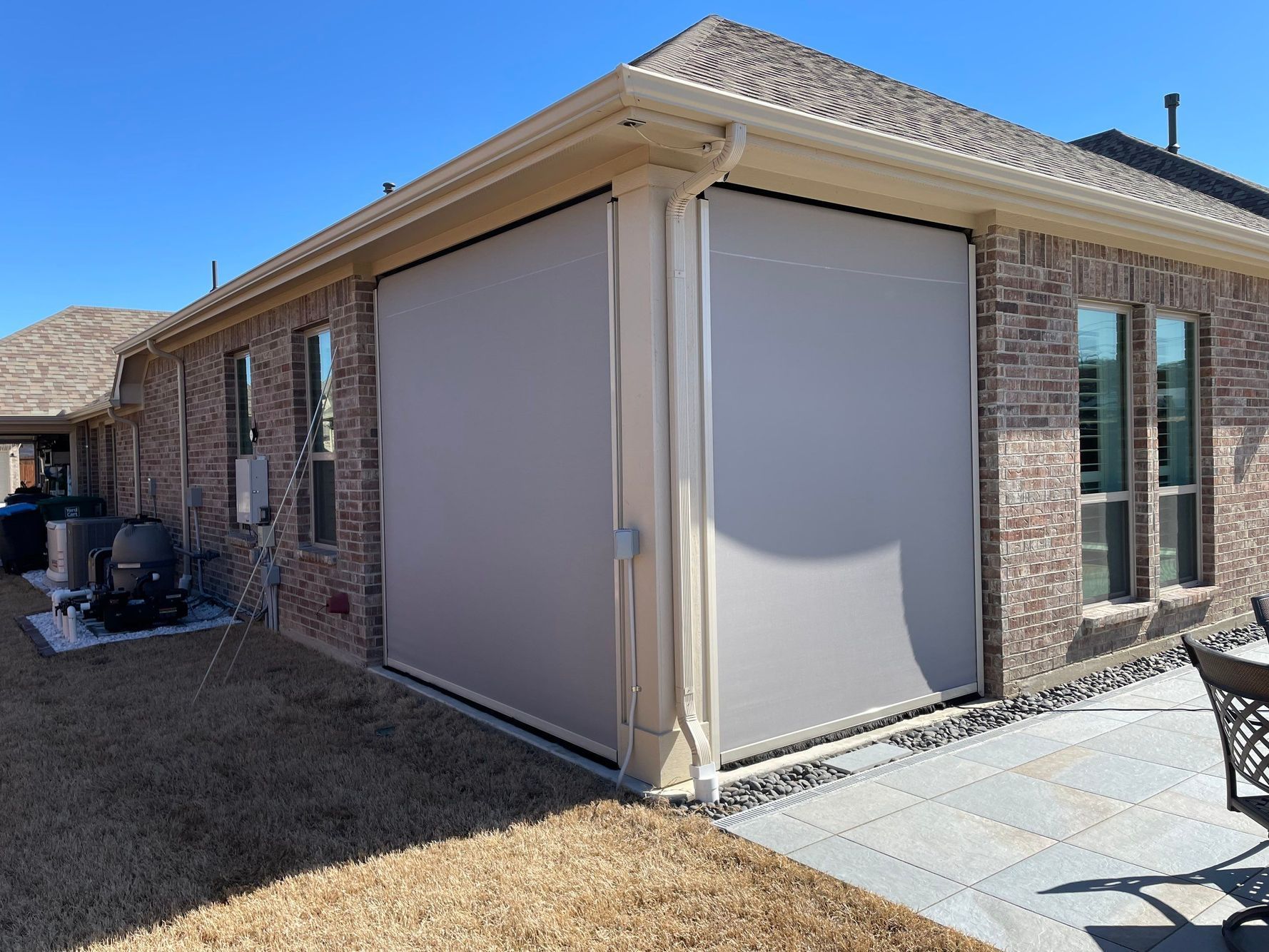 Tan retractable shades on a brick house patio, shielding the area from the sun.