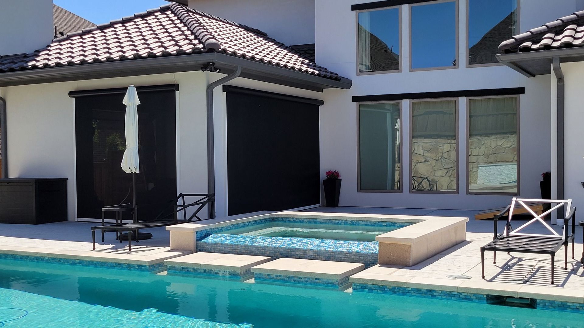 Poolside view of a white stucco house with black outdoor shades. A pool and hot tub are in the foreground.
