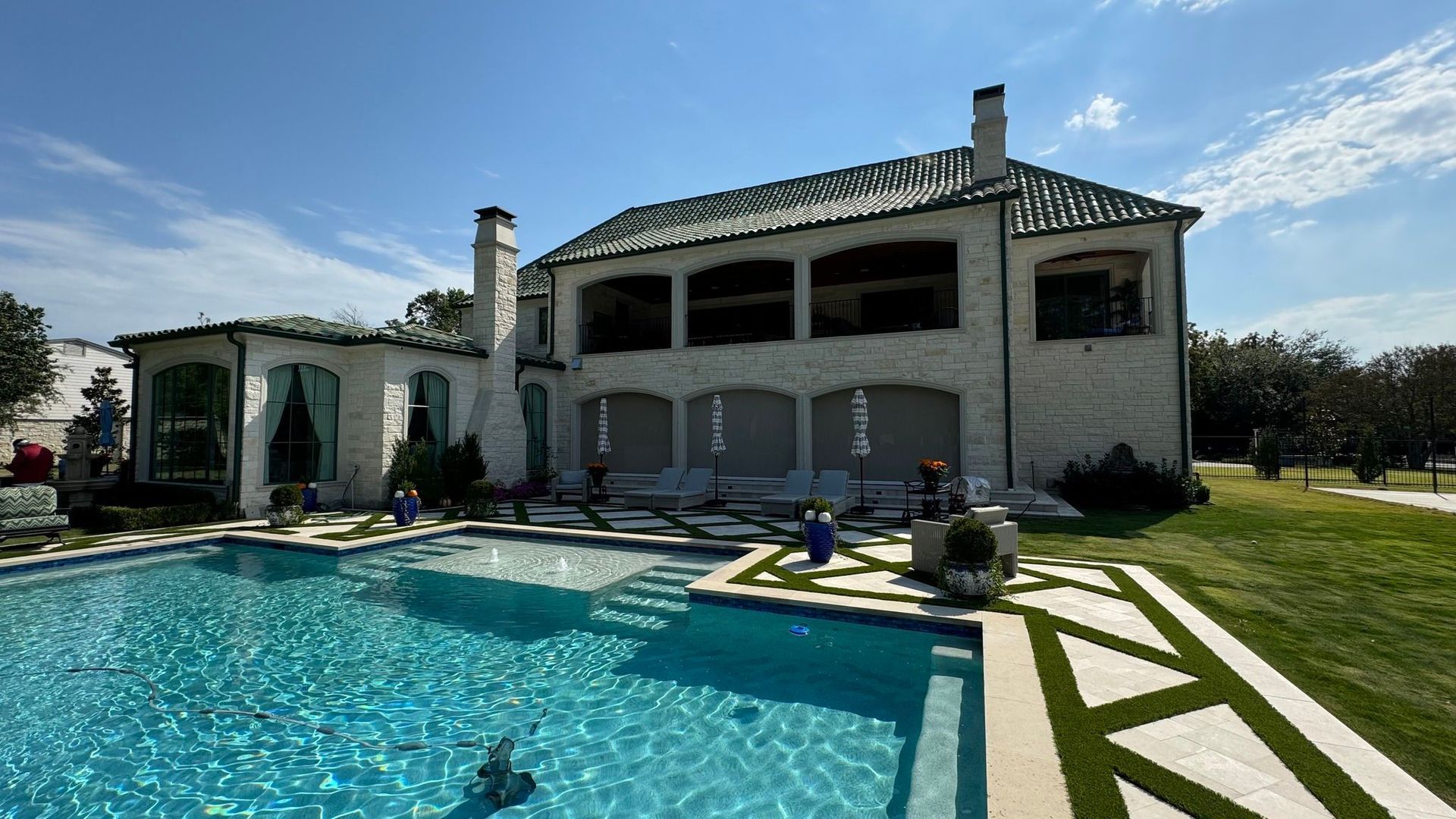 Back view of a large, white mansion with a pool, blue water, and green roof under a sunny sky.