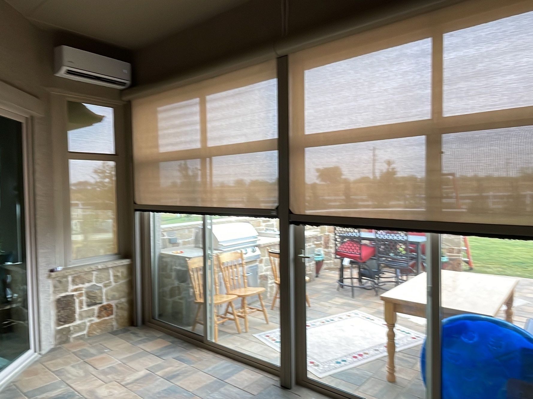Sunroom with roller shades partially covering windows, overlooking a patio with grill and table.