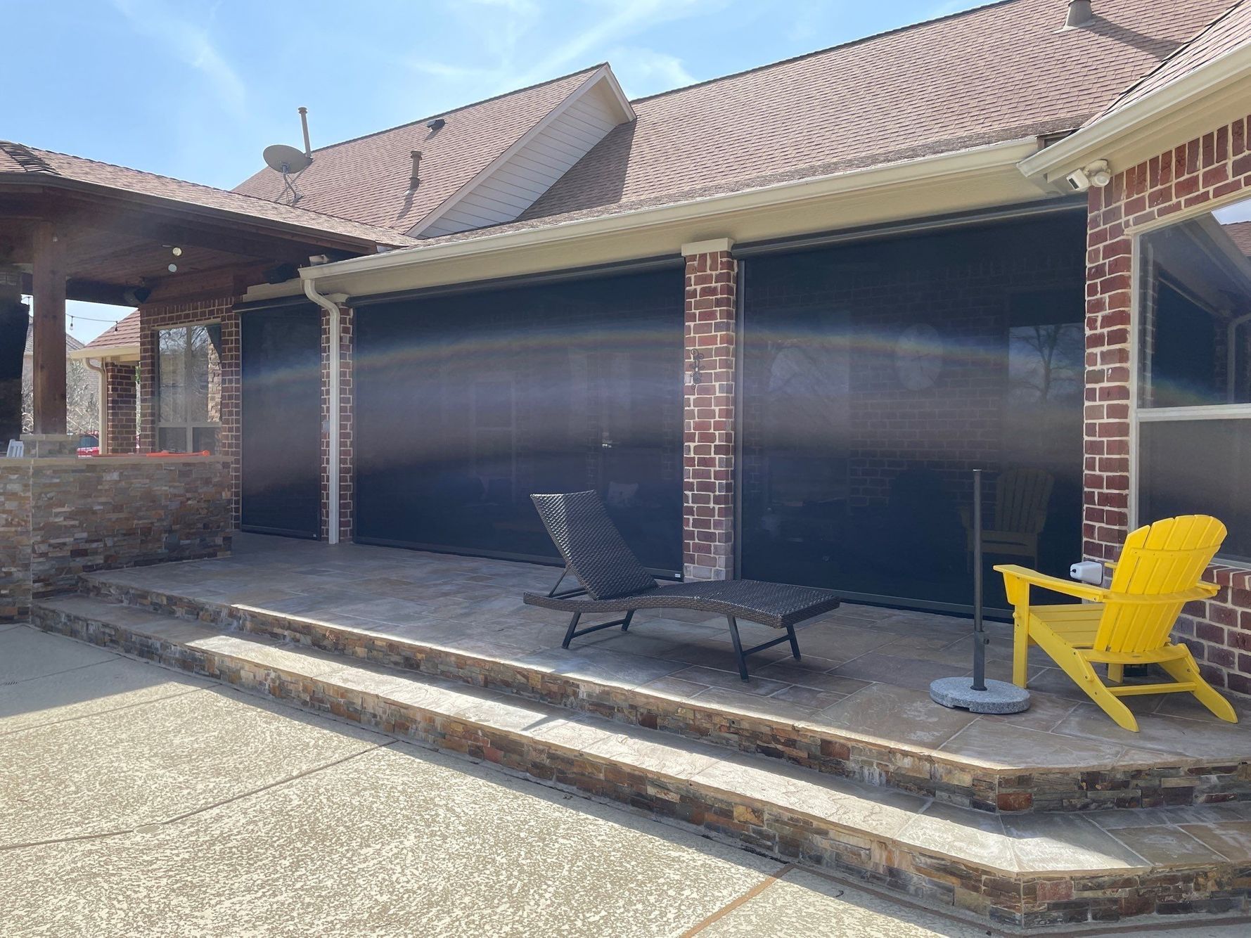 A patio with dark screens, brick columns, and a yellow chair.  A lounge chair sits in front of the screens.