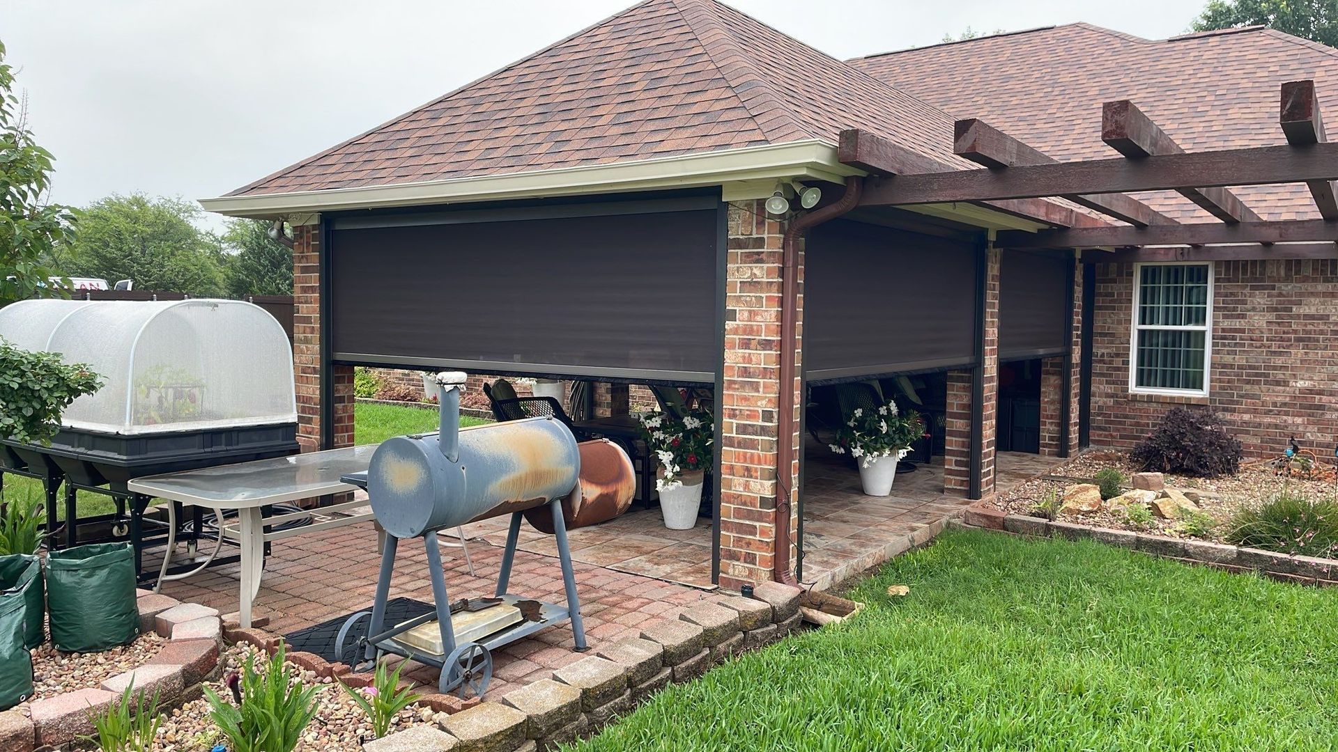 Brown outdoor shade covers a brick patio with a barbecue, table, and green lawn.