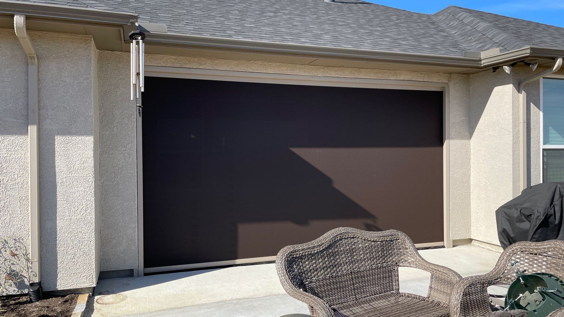 Dark brown exterior shade covers a patio opening, flanked by tan stucco walls. A wicker chair sits in the foreground.