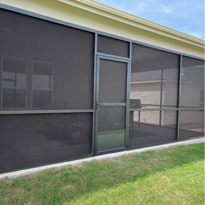 Screened-in porch with dark mesh, door, and metal frames. Green grass in the foreground, under a white building.