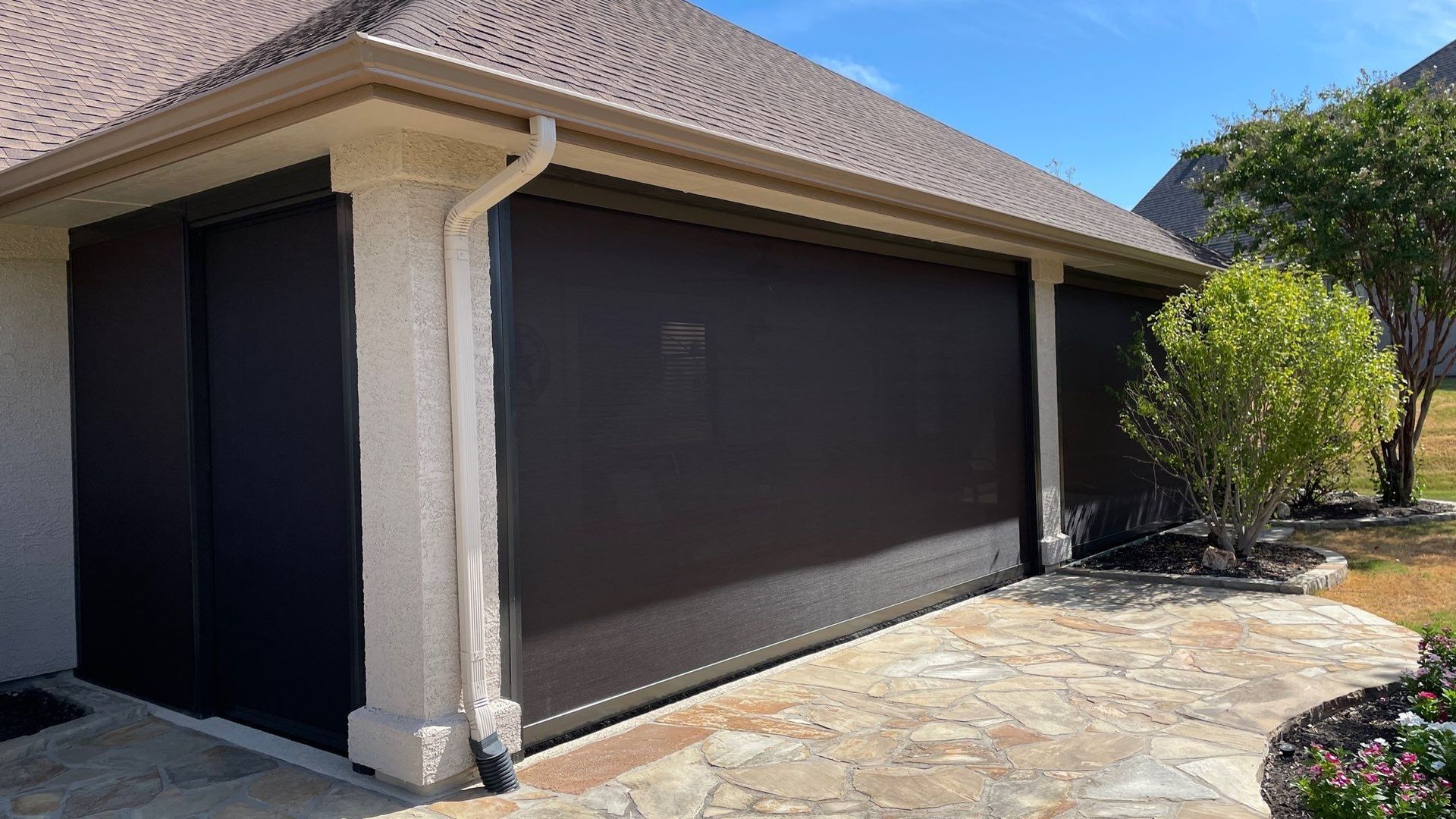 Black sun shades on a house, covering a patio. Stone path in foreground, sunny day.