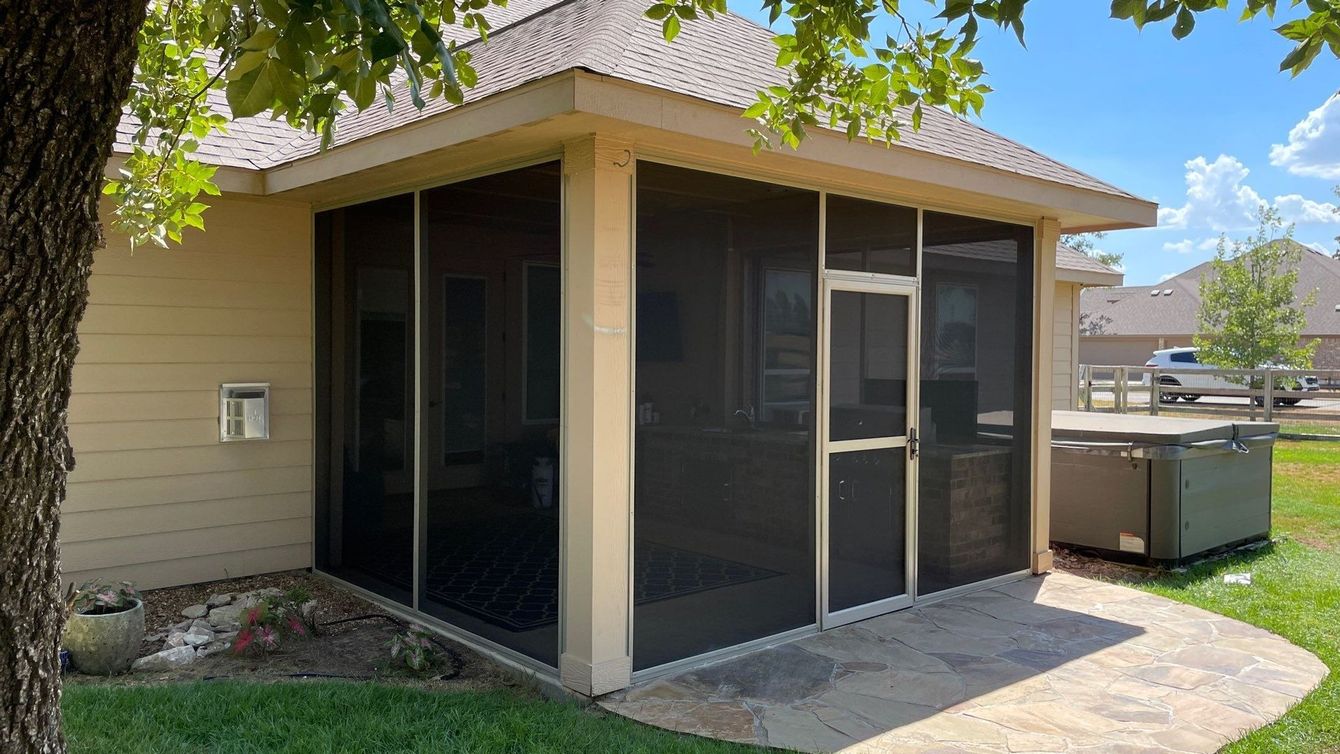 Screened-in patio extension attached to a house. Beige walls, dark screens, concrete patio, and small hot tub visible.