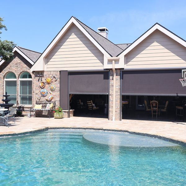 Backyard pool in front of a beige house with open garage with privacy screens and stone-accented exterior.