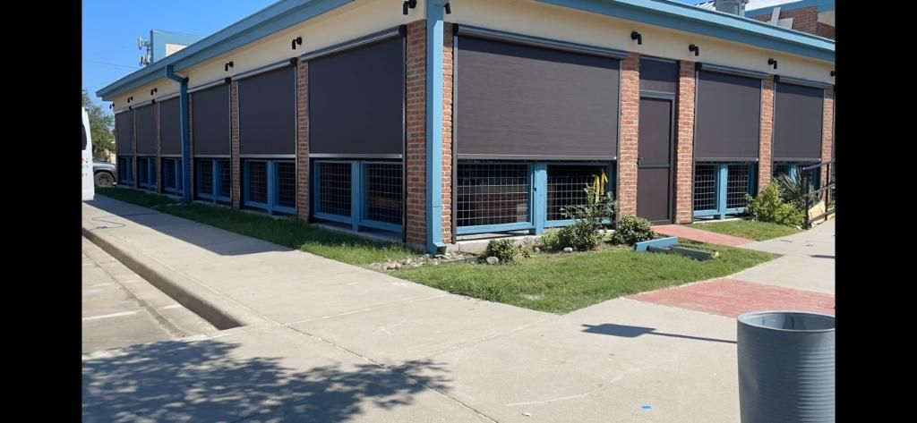 A building with dark window coverings, brick accents, and a blue trim. A sidewalk and grass border the building.
