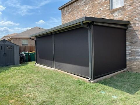 Dark gray outdoor shades enclose a patio attached to a brick house. Green lawn and a small shed in background.