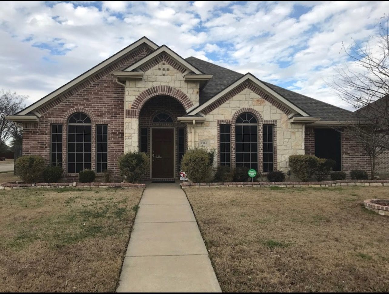 House with red brick and beige stone exterior, arched doorway, and windows. Brown lawn, cloudy sky.