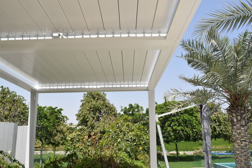 White pergola with adjustable slats, overlooking lush greenery and a palm tree under a blue sky.