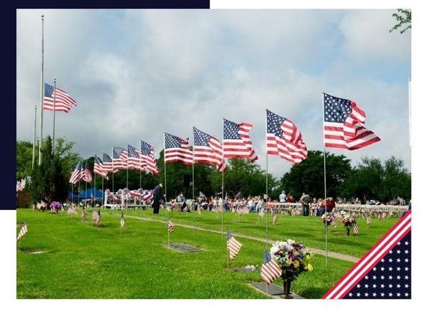 American flags at a cemetery, many at half-staff, people gathered, sunny day.
