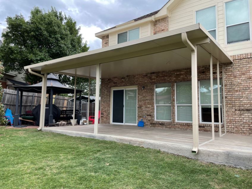 Backyard patio with beige roof, columns, and brick wall. Green grass and gazebo in the background.