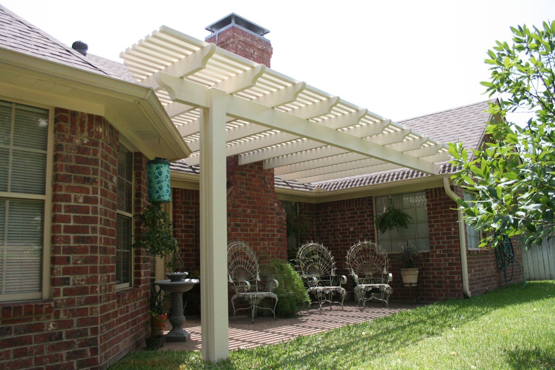 White pergola attached to a brick house, over a brick patio with chairs.