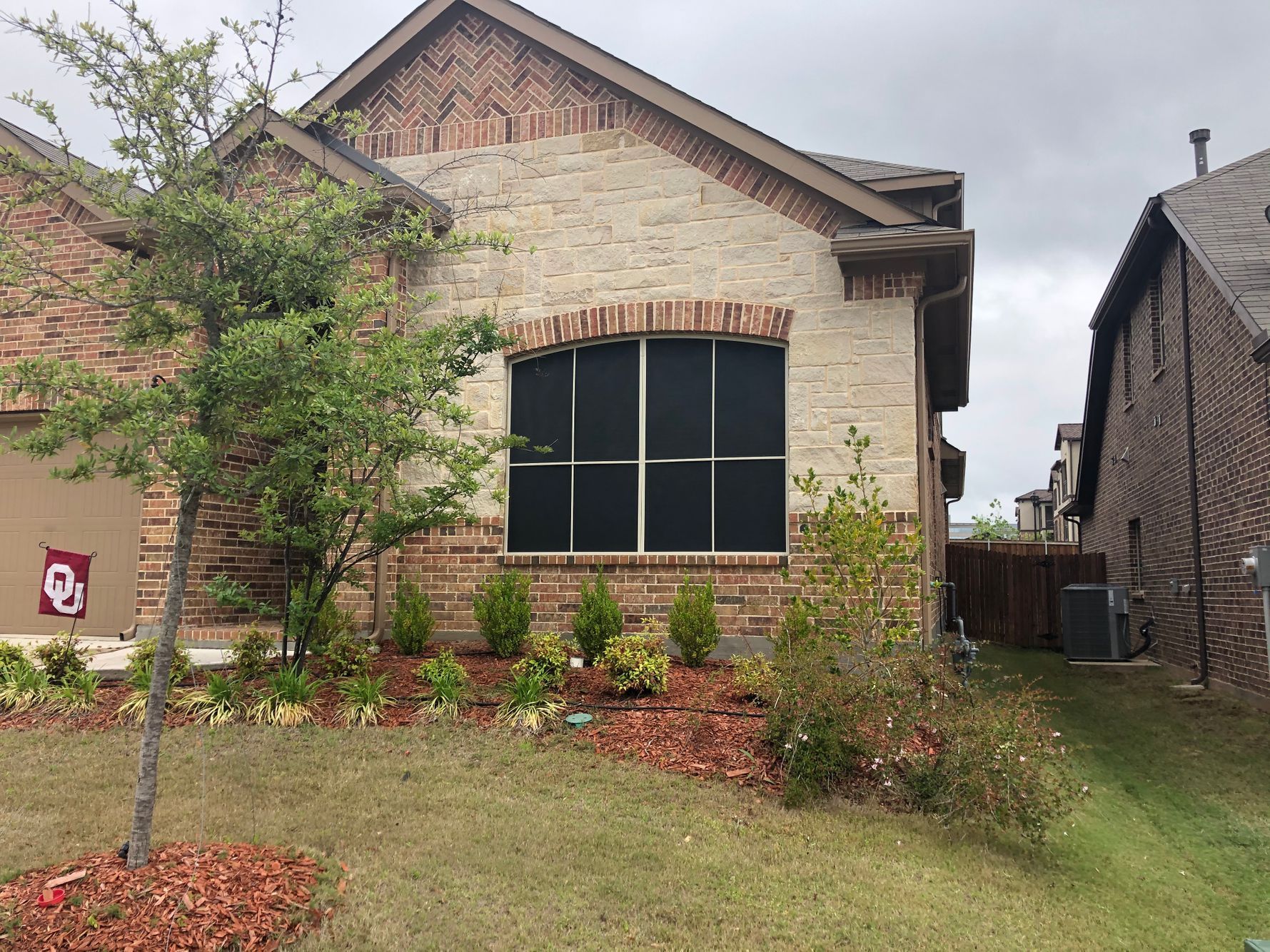 Brick house with arched window covered by black screen, small bushes in front, overcast sky.