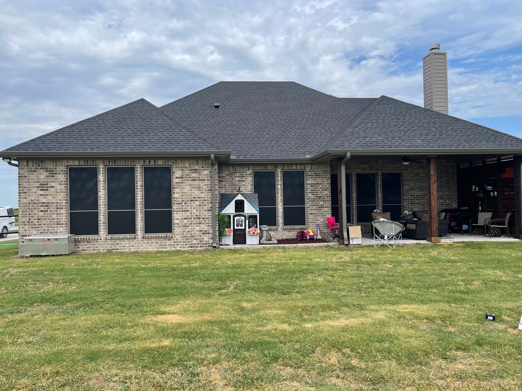 Back of a brick house with a dark roof, windows, and a covered patio. A playhouse is in the yard.