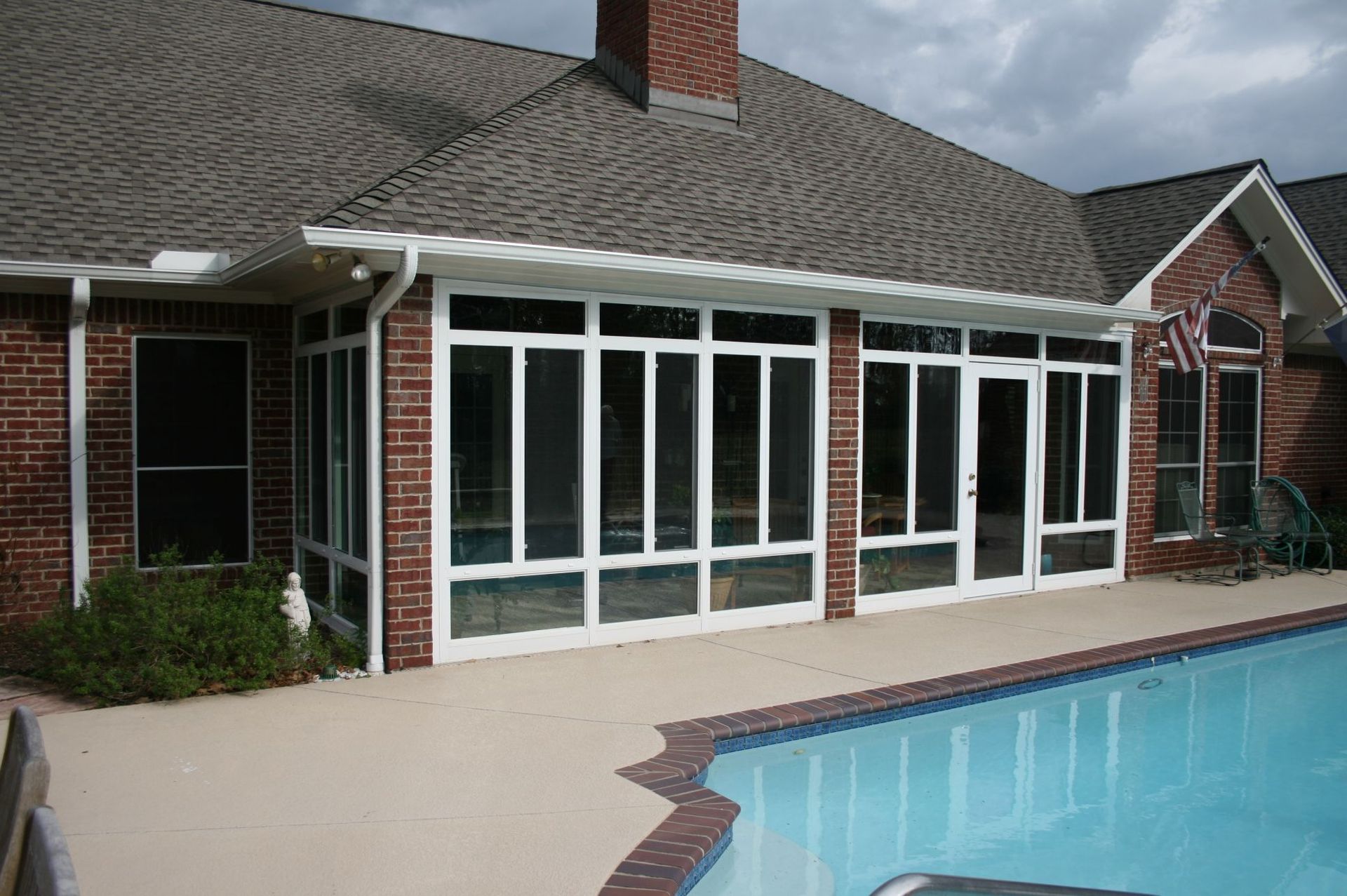 Screened-in porch attached to a brick house with pool. White frames, glass panels, and brick chimney visible.