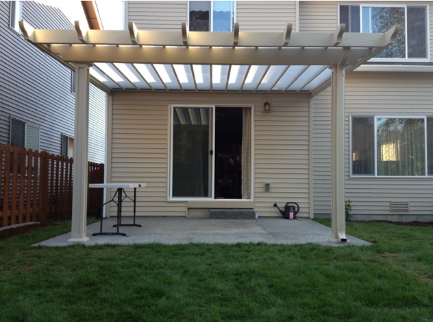 Beige house with sliding glass door and concrete patio, covered by a white pergola, on green lawn.