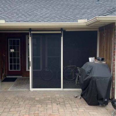 Screen door on a patio, white frame, black screen, next to a covered grill. Brick flooring.