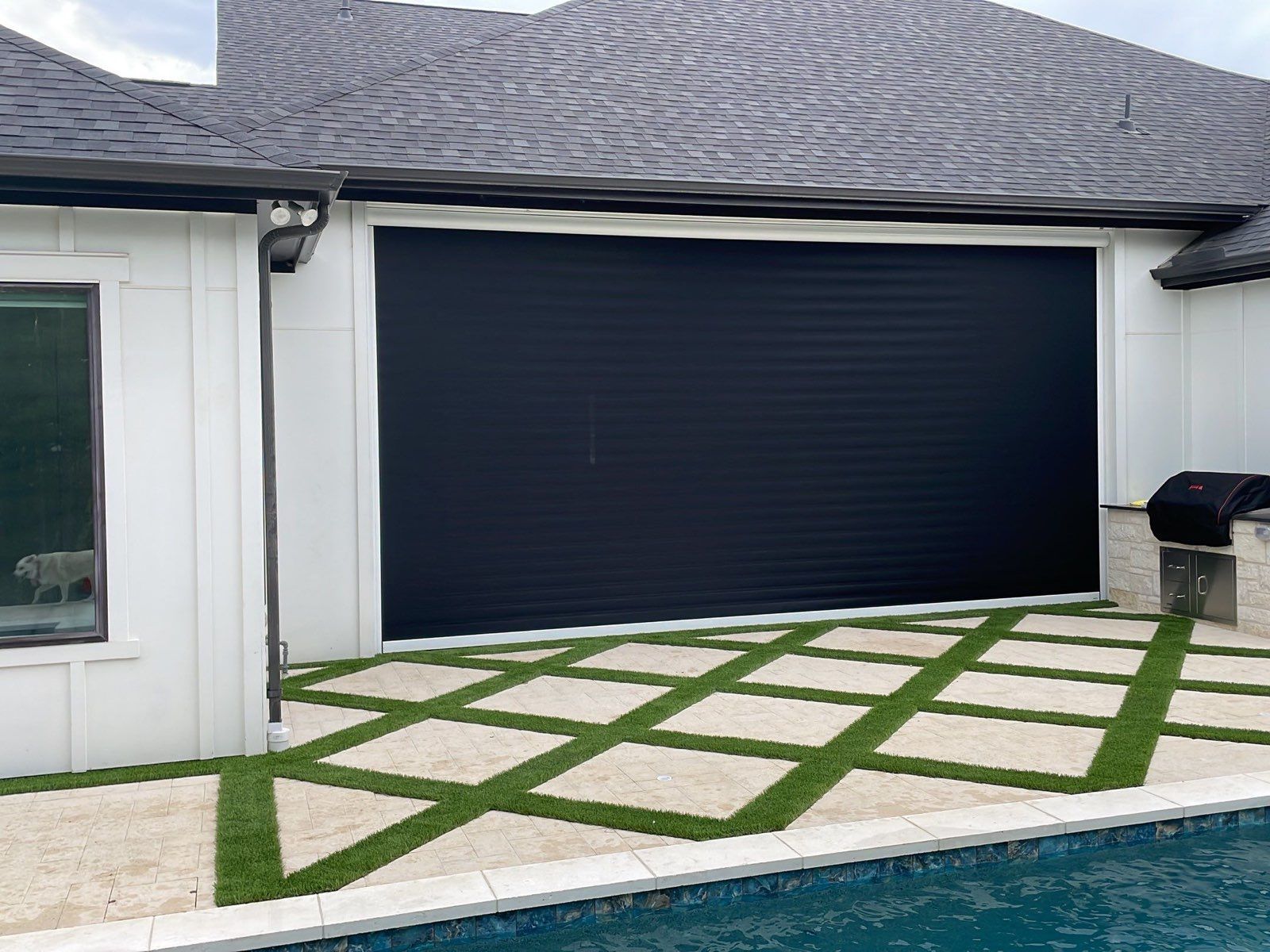 Black retractable screen on a white house, covering a patio. Lawn and stone patio with a pool.