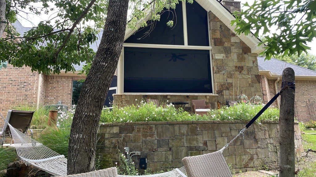 Exterior of a stone house with a hammock in the foreground. Large arched window with black shade. Lush greenery.