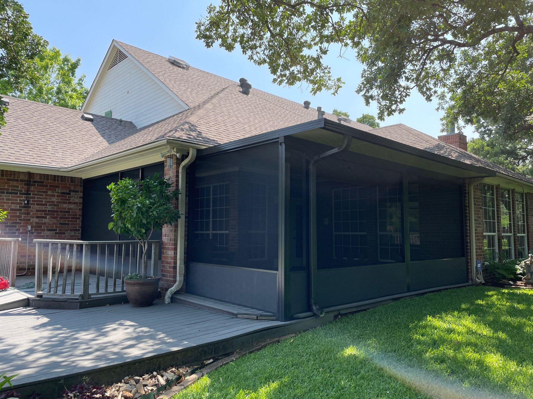 Screened-in porch attached to a brick house with dark screens and a gray roof. Green grass and trees surround it.