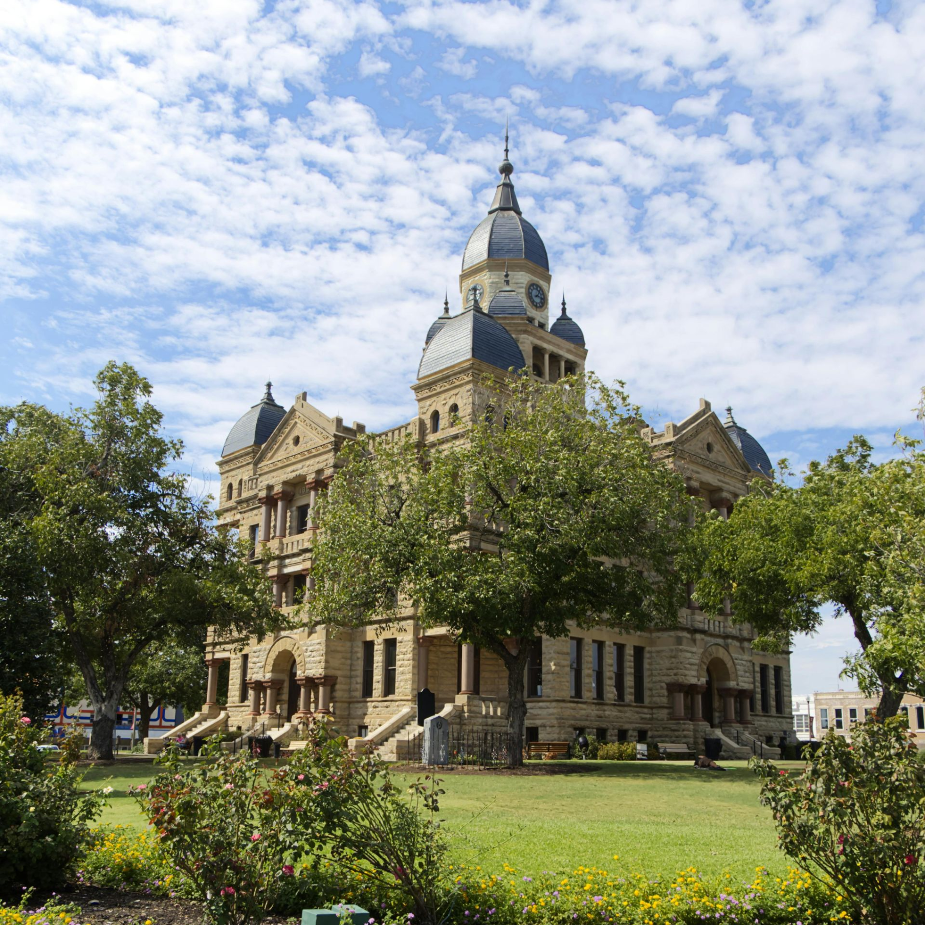 Historic courthouse with a clock tower under a cloudy sky, surrounded by trees and a flowerbed.